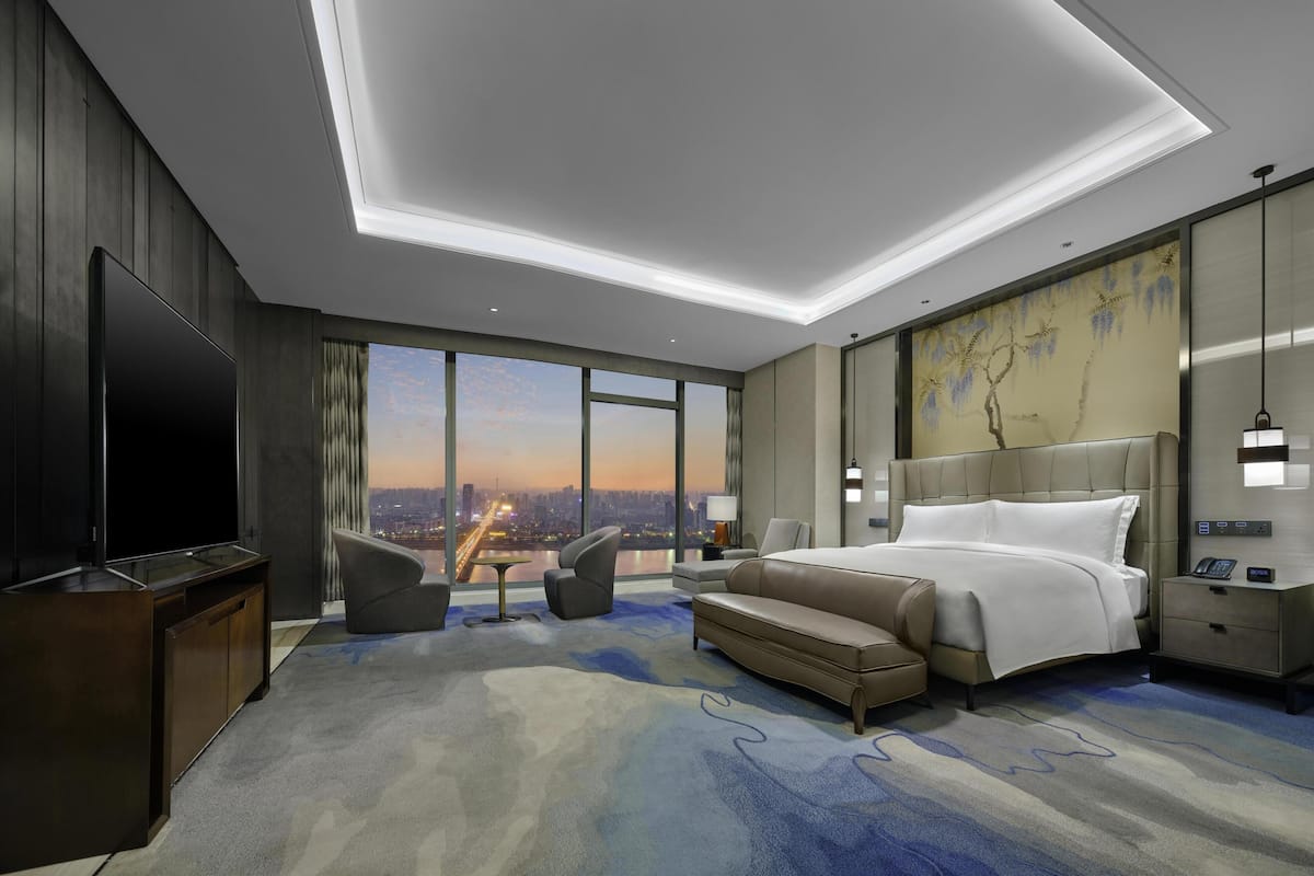 A modern hotel room with a large bed, a TV, a desk, and a view of the cityscape.