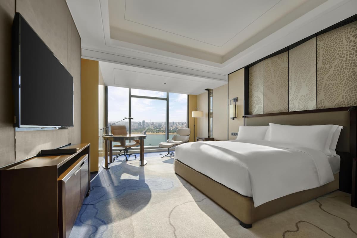 A hotel room with a large bed, a desk with a chair, a TV, and a view of a cityscape through large windows.
