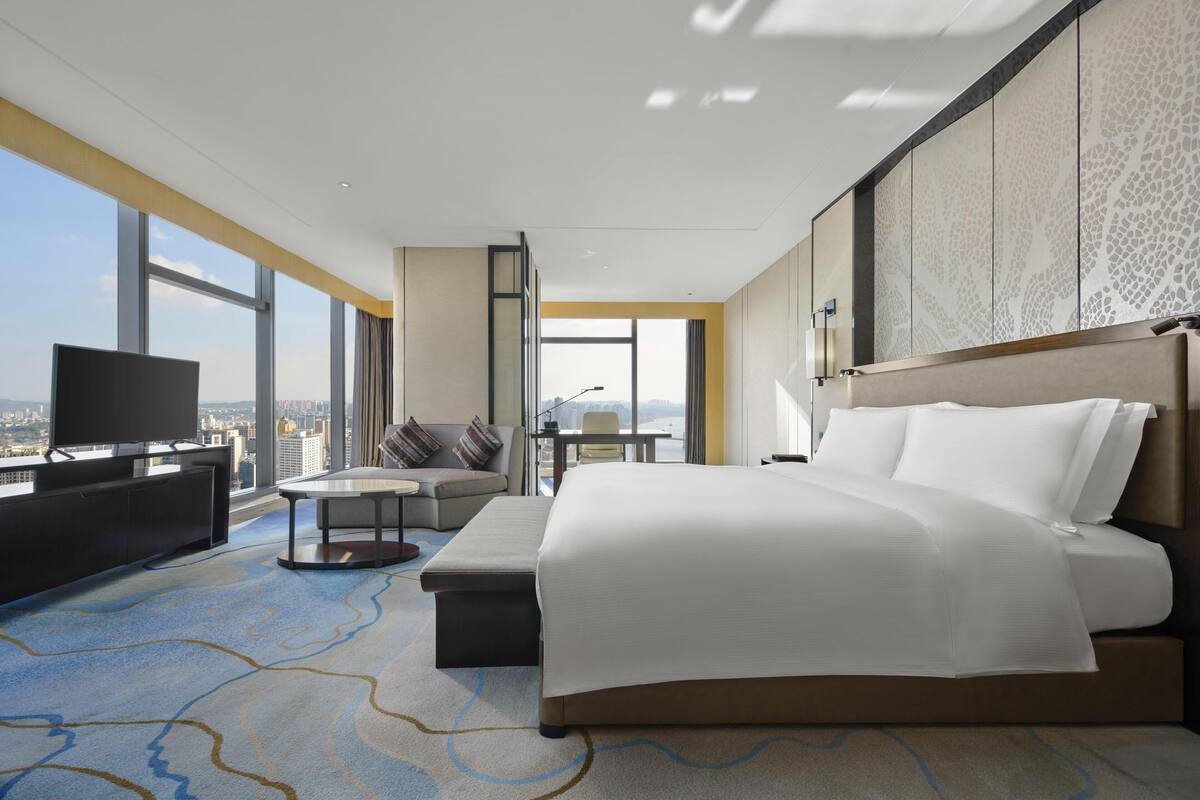 A modern hotel room with a large bed, a TV, a seating area, and a view of the city.