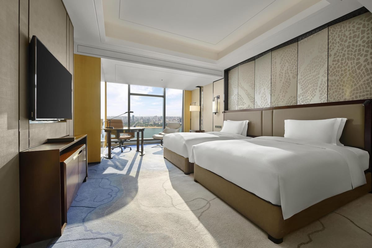 A hotel room with two beds, a desk, a TV, and a large window with a city view.