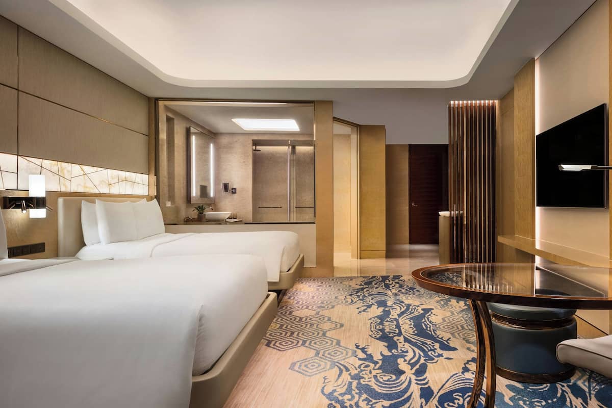 A modern hotel room with two beds, a dining table, a TV, and a bathroom visible through an open door.