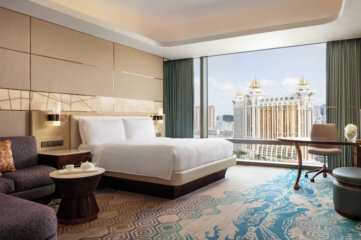 A modern hotel room with a large bed, a sitting area with a sofa and armchair, a small table with a teapot, and a view of a cityscape through large windows.