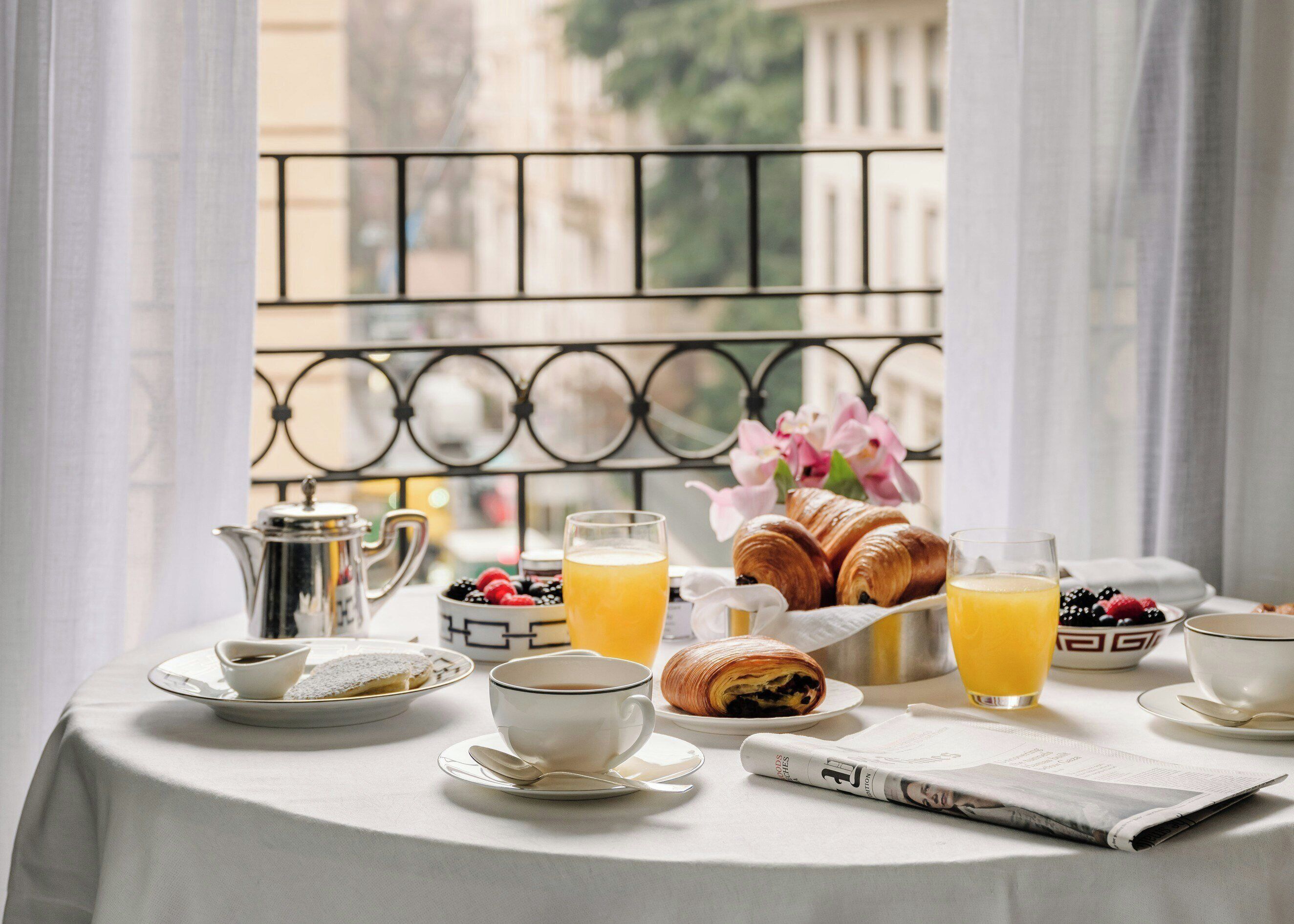 A breakfast table with croissants, fruit, coffee, and orange juice, set on a balcony with a view of buildings.
