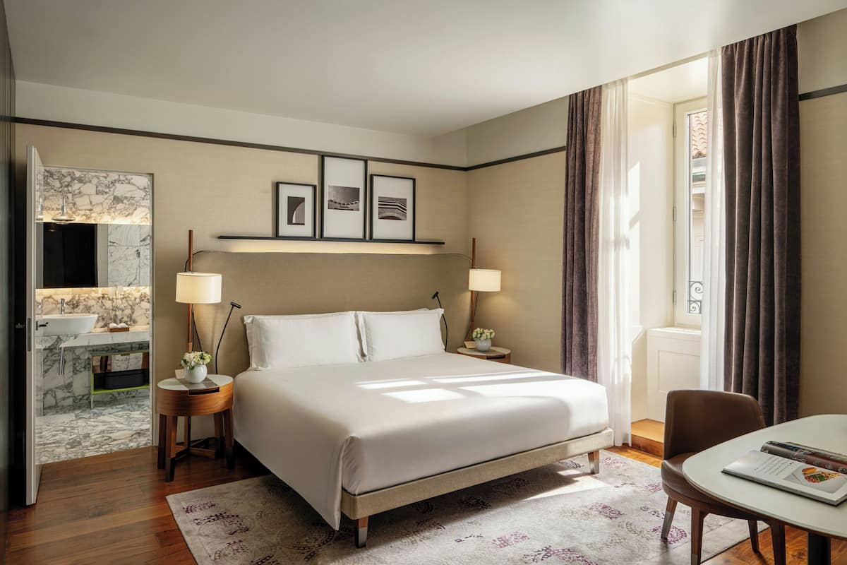 A hotel room with a large bed, bedside lamps, a small table with a magazine, a bathroom with a marble sink, and framed pictures on the wall.