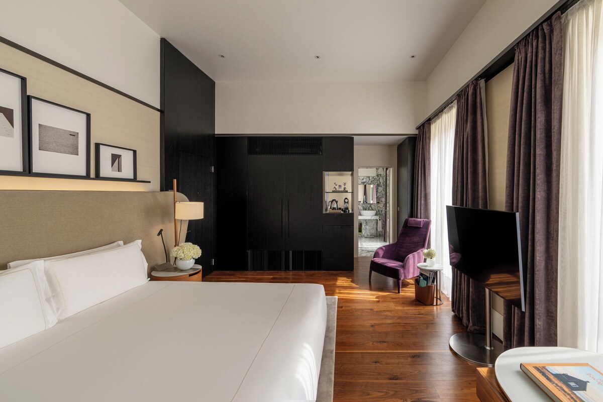 A modern hotel room with a large bed, a flat-screen TV, a seating area with a purple armchair, and a small table with a lamp and flowers.