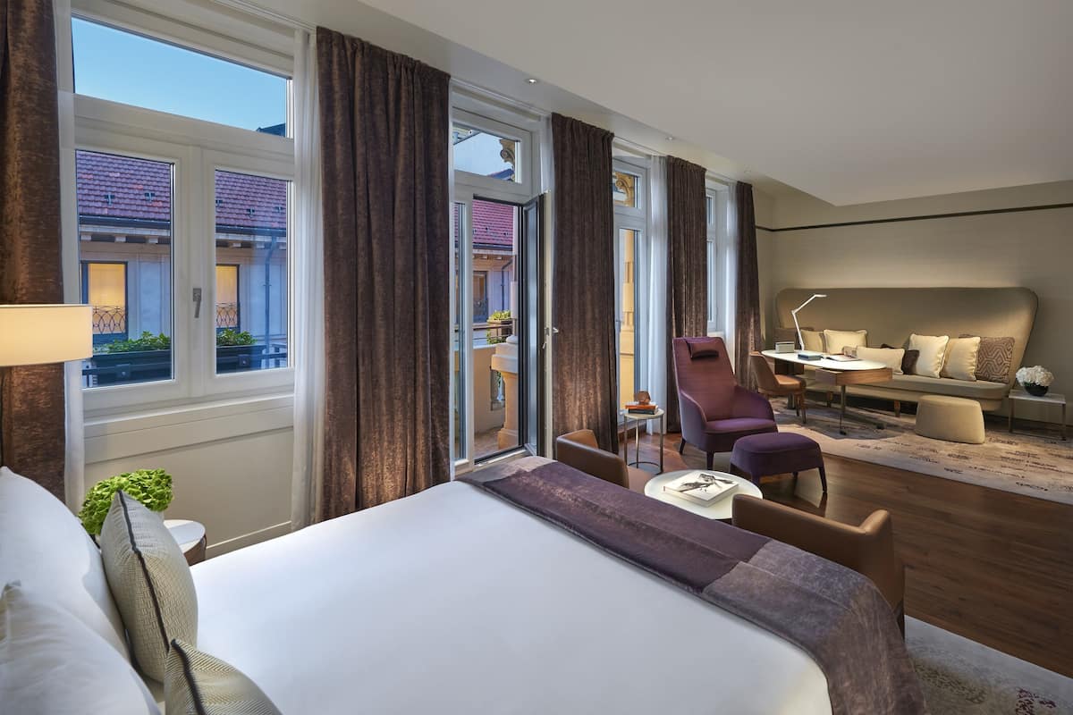 A hotel room with a large bed, a desk, a chair, and a sofa. There is a window with curtains, a plant, and a view of the outside.