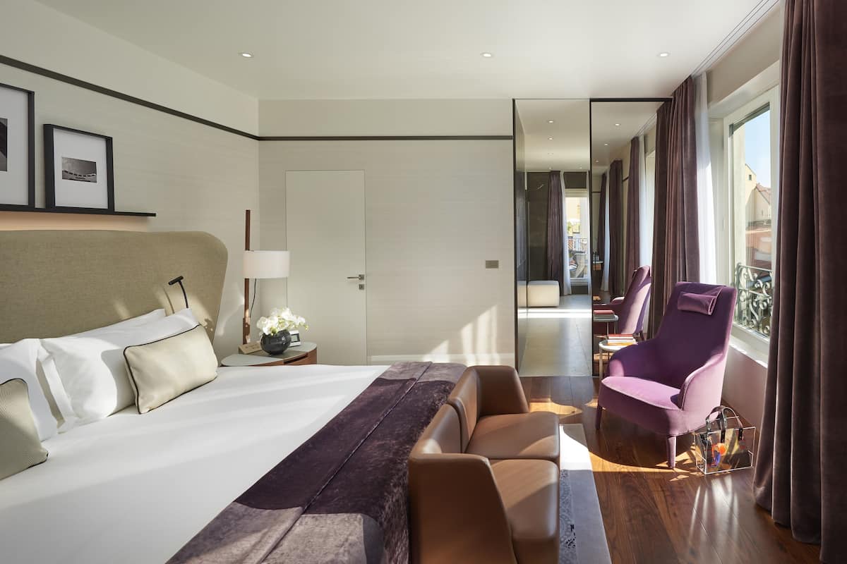 A hotel room with a large bed, a sitting area with a purple chair, a small table with a lamp, and a view of the outside balcony.