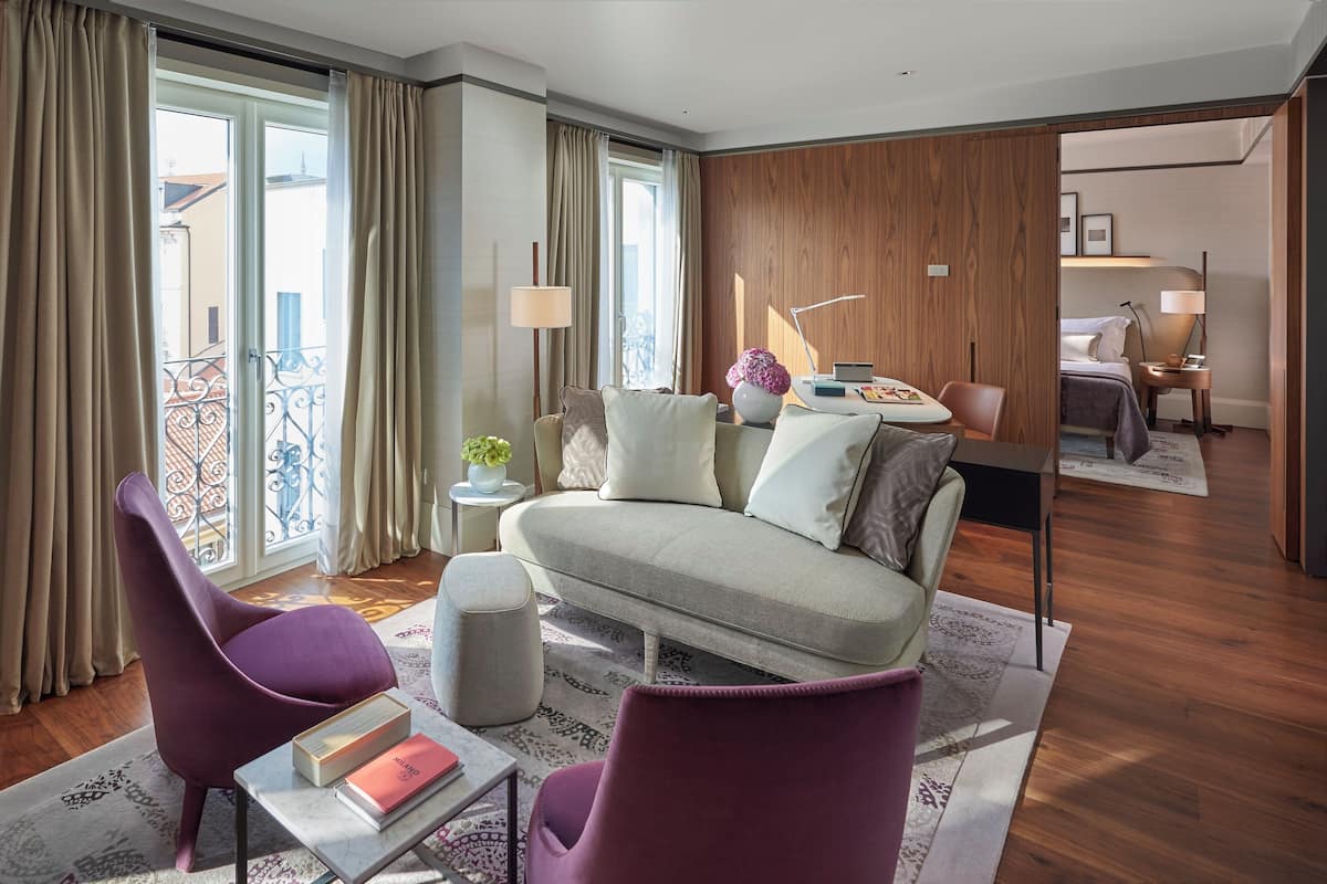 A modern hotel room with a sofa, two armchairs, a small side table, and a bed.