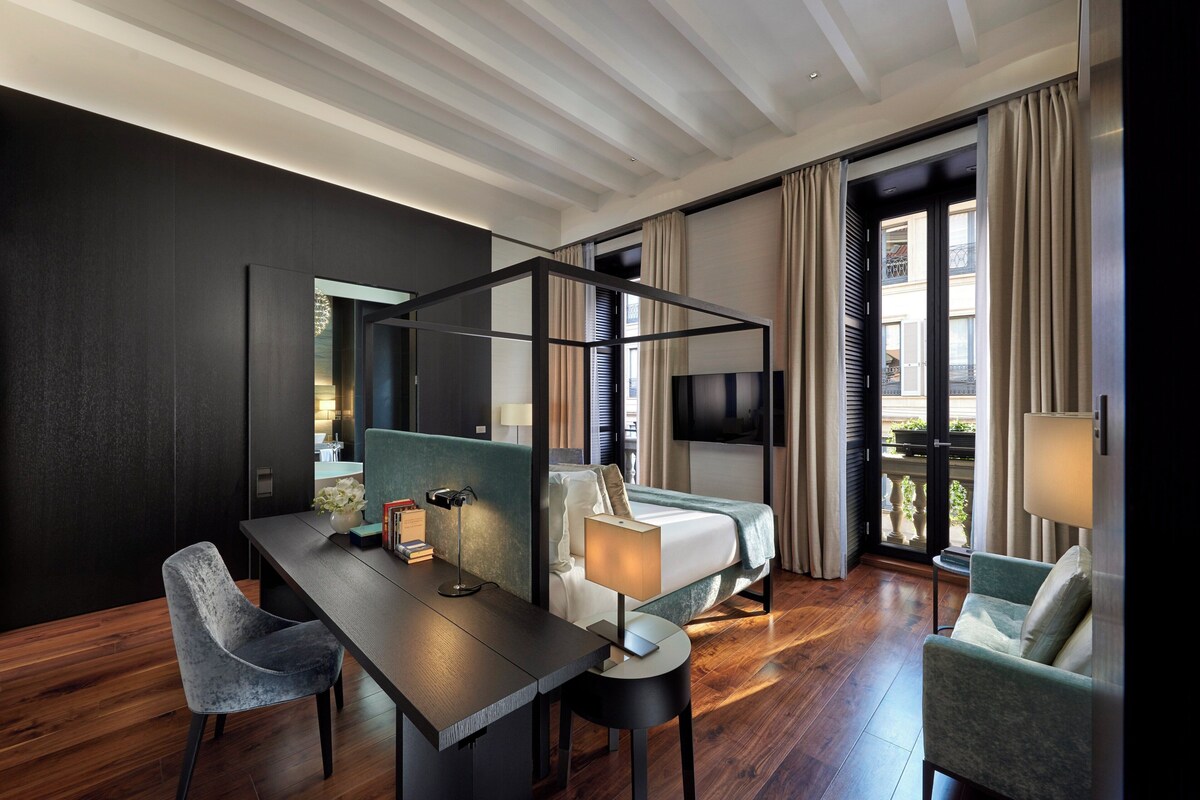 A modern hotel room with a bed, a desk, a chair, a TV, and a balcony with curtains.