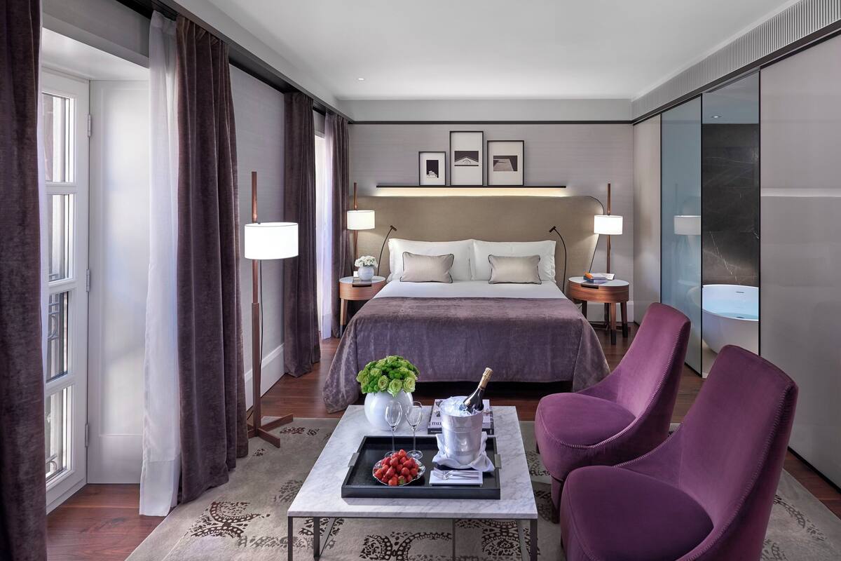 A hotel room with a bed, bedside tables, a coffee table with a tray of strawberries, a vase of flowers, and a champagne bottle.