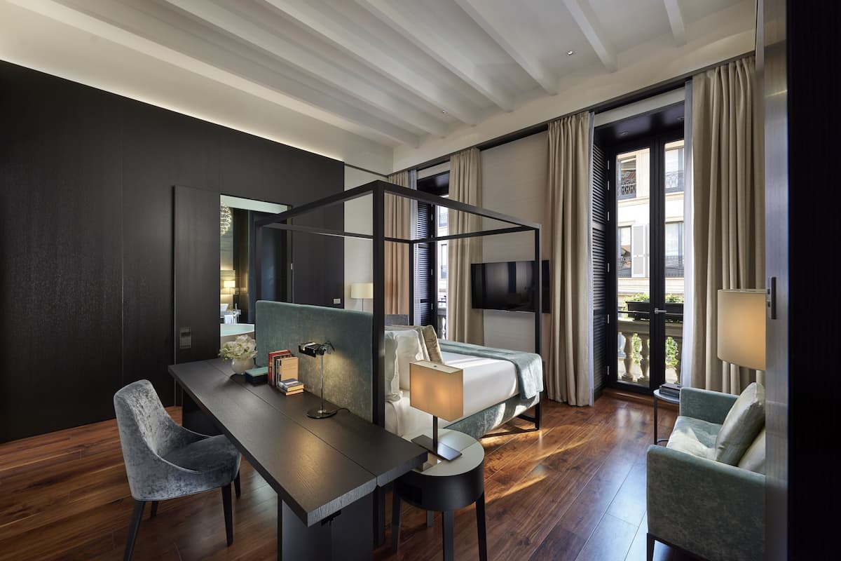 A modern hotel room with a bed, a desk, a chair, a TV, and a balcony with curtains.