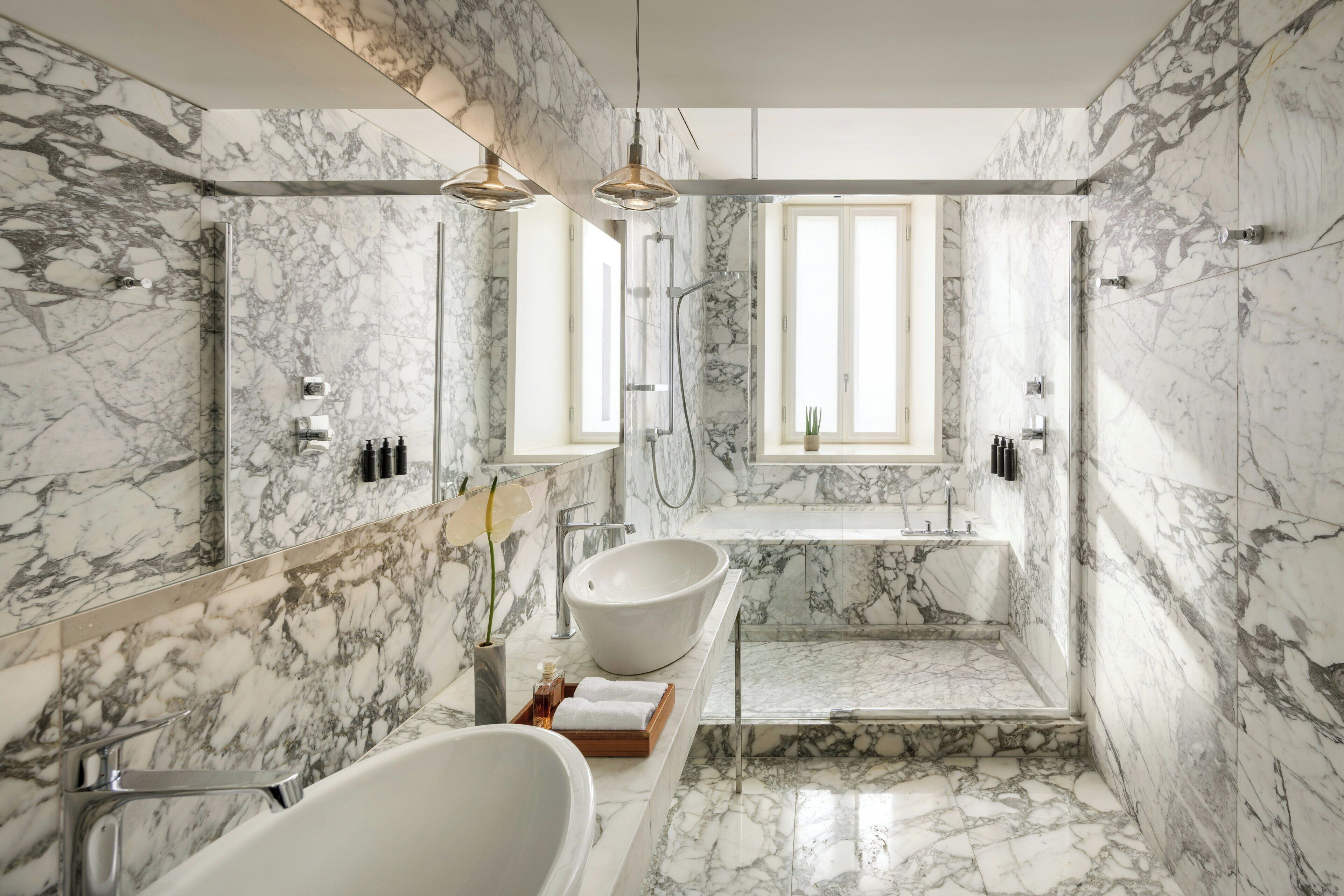 Bagno con parete in marmo, vasca freestanding, lavabo a incasso, zona doccia e illuminazione a sospensione.