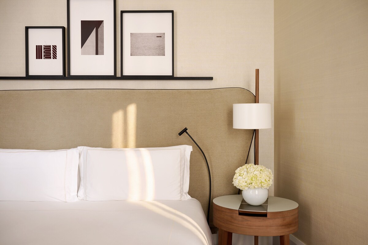 A neatly made bed with white linens, a bedside table with a lamp and a vase of flowers, and a wall with three framed pictures.