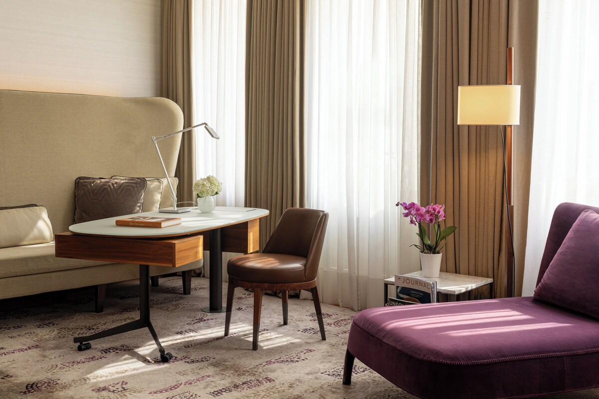 A hotel room with a sofa, a chair, a small table with a lamp, a potted plant, and a purple armchair.
