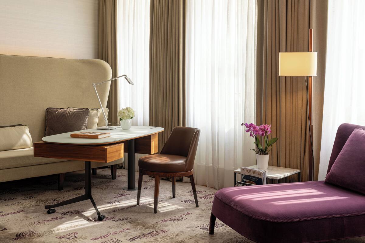 A hotel room with a sofa, a chair, a small table with a lamp, a potted plant, and a purple armchair.