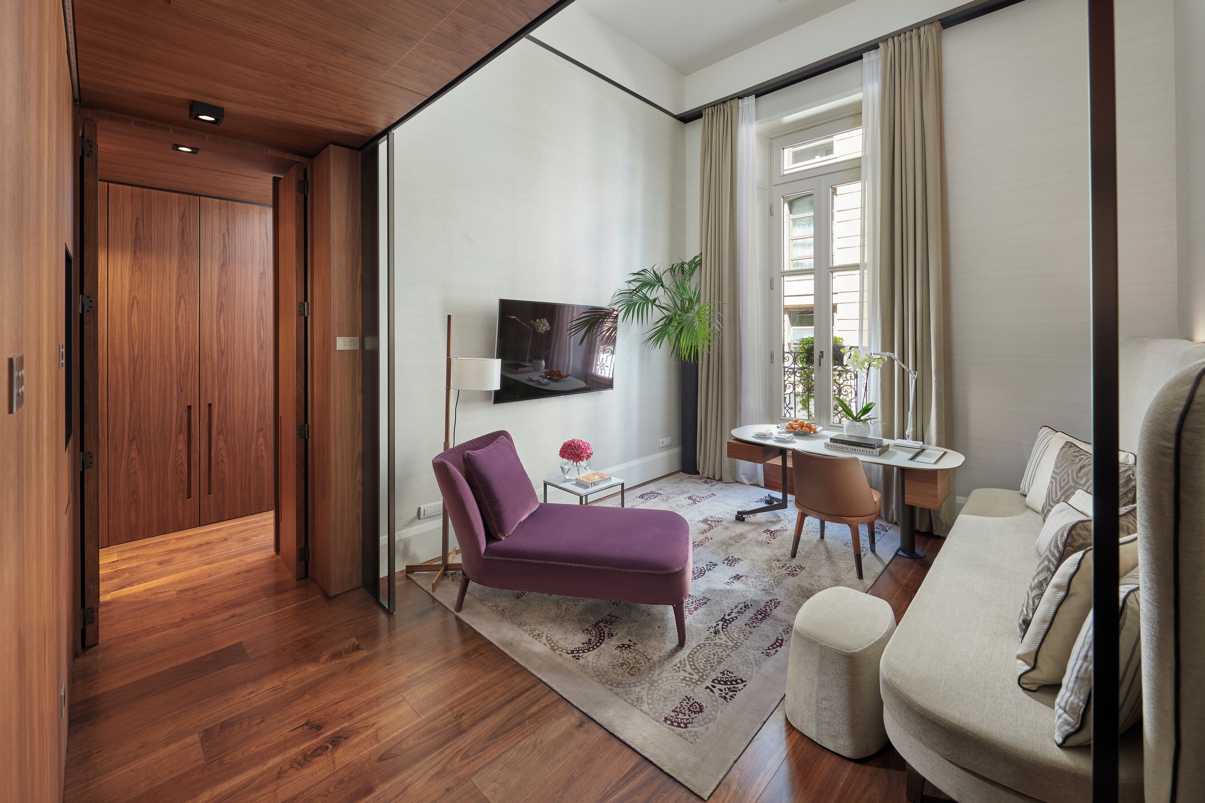 Una camera d'albergo moderna con pavimento in legno, una poltrona viola, un divano, un tavolino con una pianta e una TV a parete.