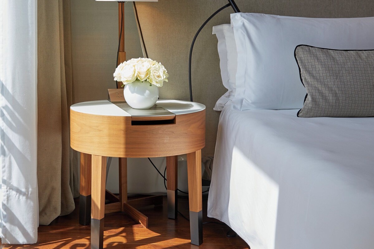 Ett hotellrum med en säng, ett nattduksbord med en lampa, en vas med blommor och en matta.