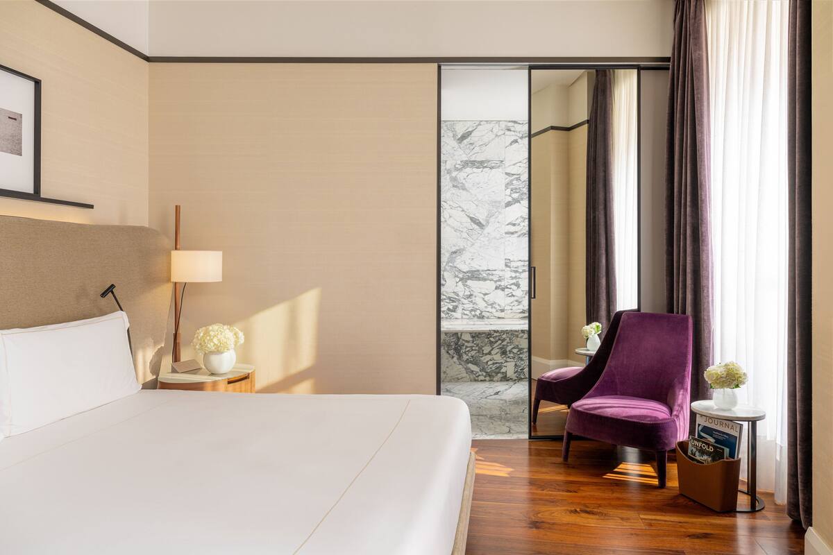 A hotel room with a large bed, a nightstand with a lamp and flowers, a purple armchair, a small table with a magazine, and a marble wall.