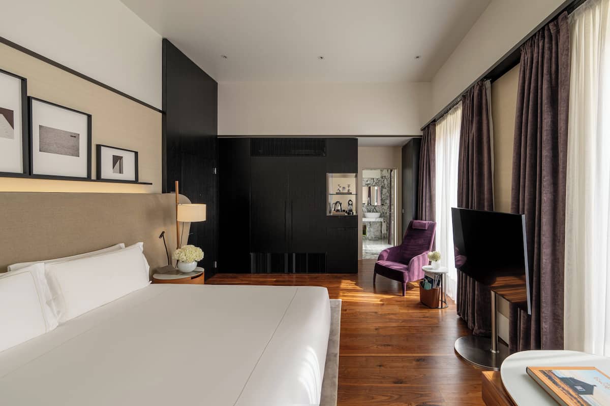 A modern hotel room with a large bed, a flat-screen TV, a seating area with a purple armchair, and a small table with a lamp and flowers.