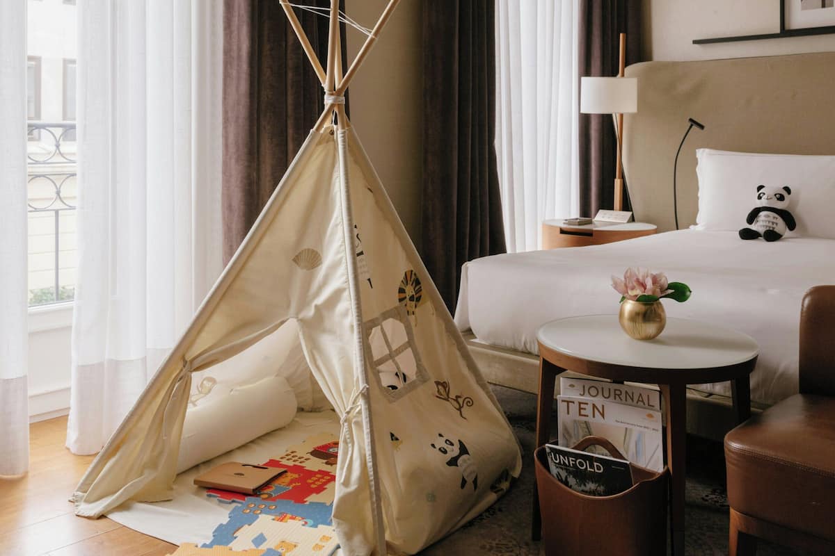 A hotel room with a bed, a small table with a lamp, a chair, a teepee, and a rug.
