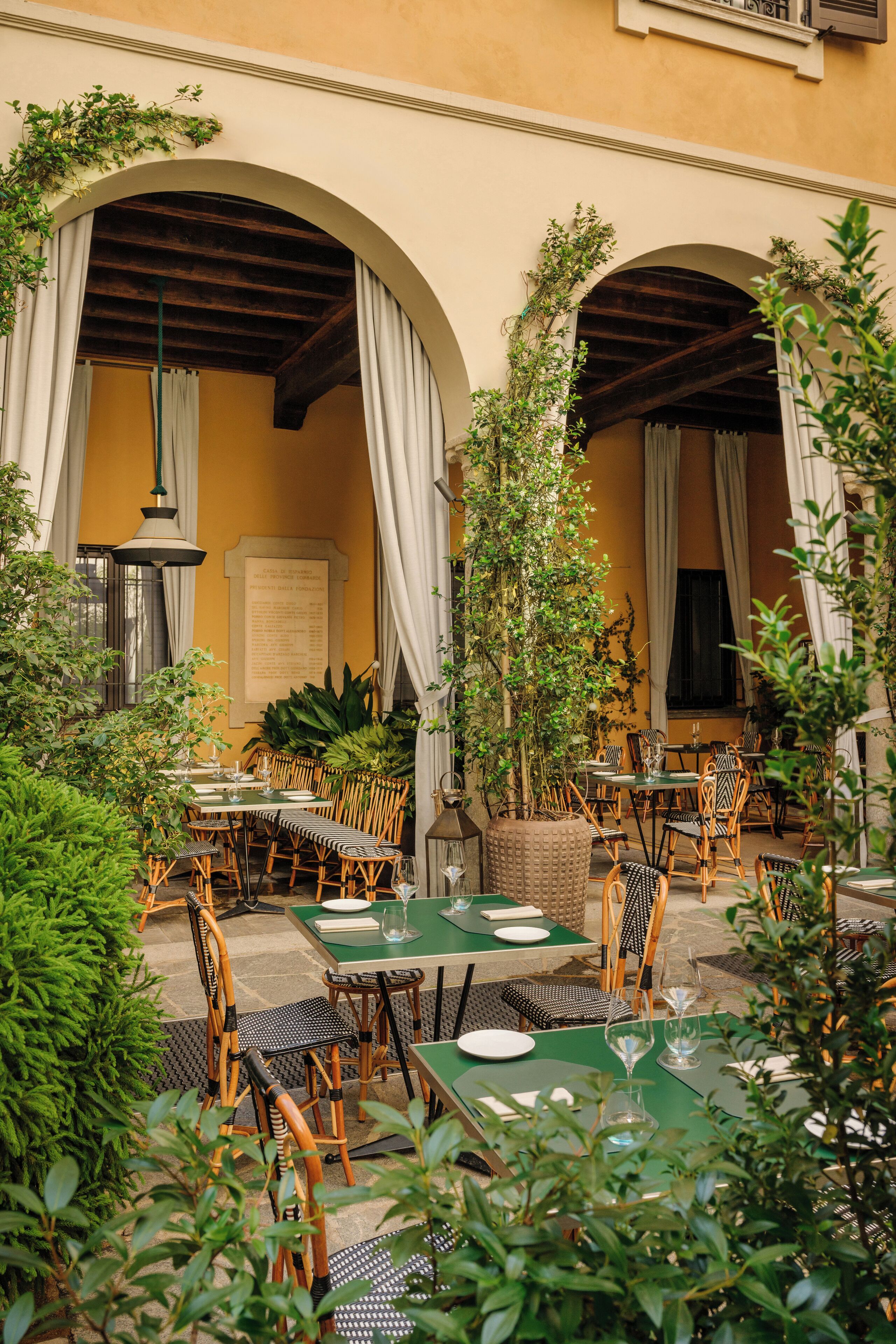 Ein Straßencafé mit Tischen und Stühlen, umgeben von Grünpflanzen und architektonischen Bögen.