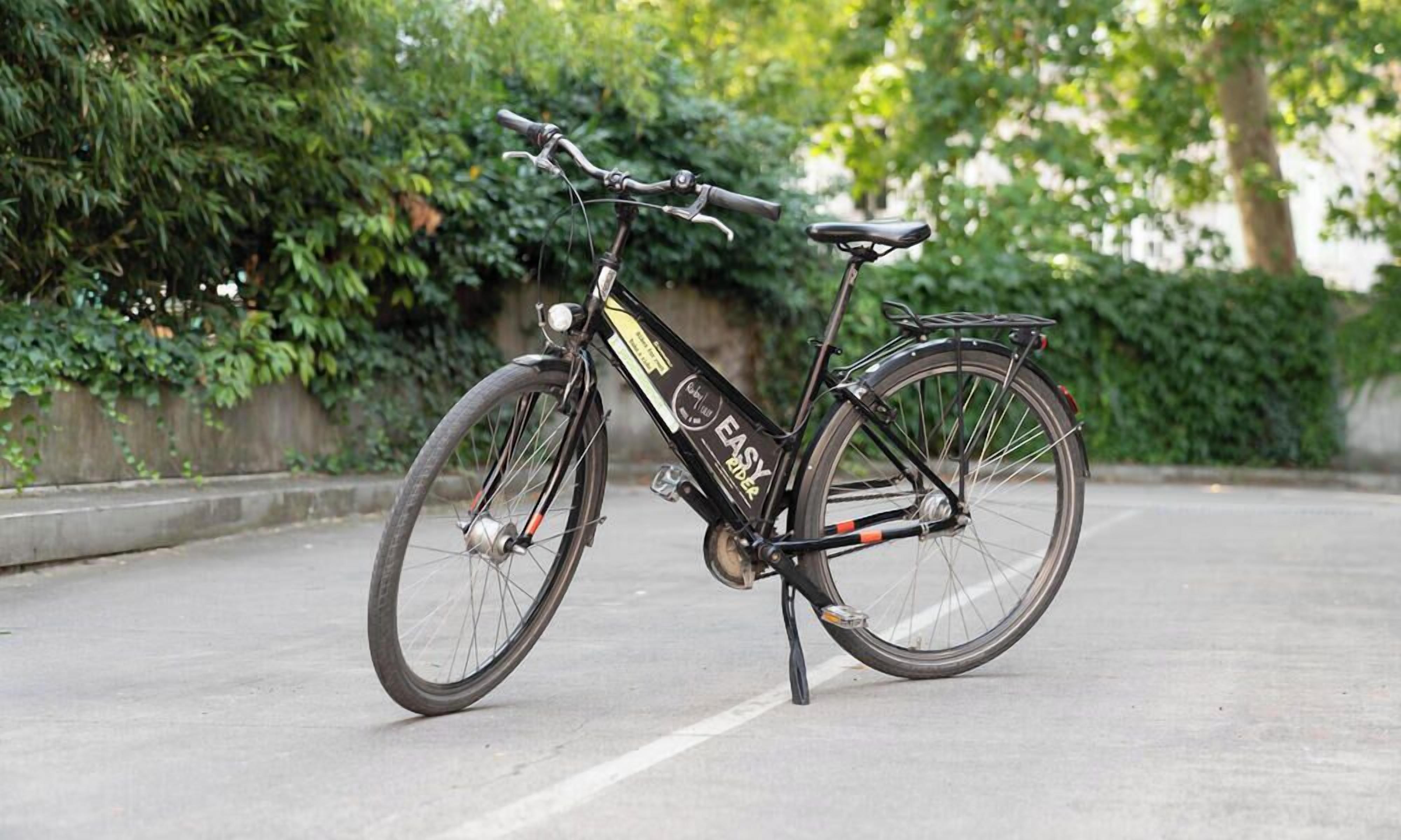 A stationary bicycle with a black frame and a branded label, positioned on a paved surface with greenery in the background.
