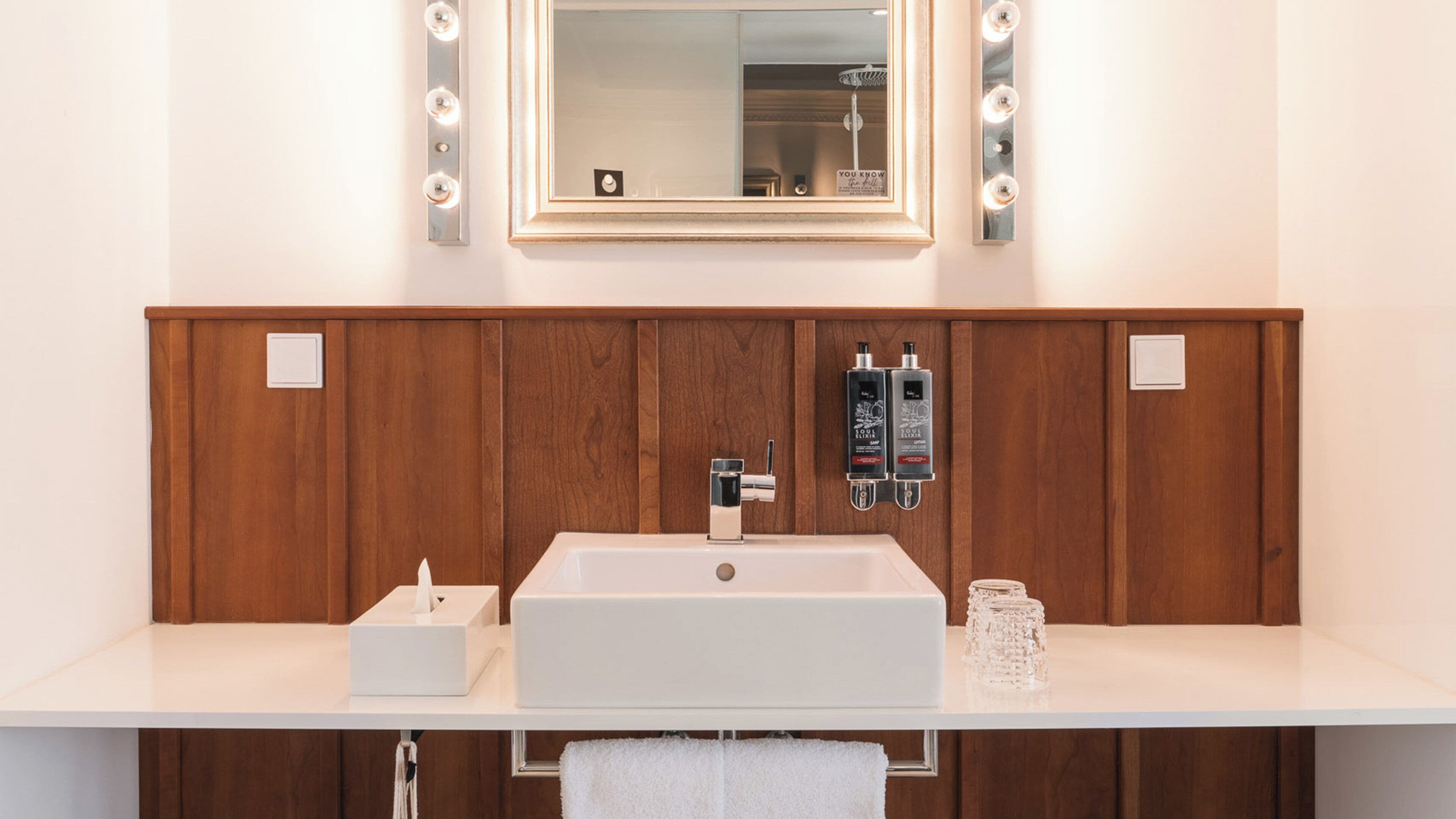 A bathroom with a wooden vanity, a mirror with lights, a sink, and two bottles of lotion.