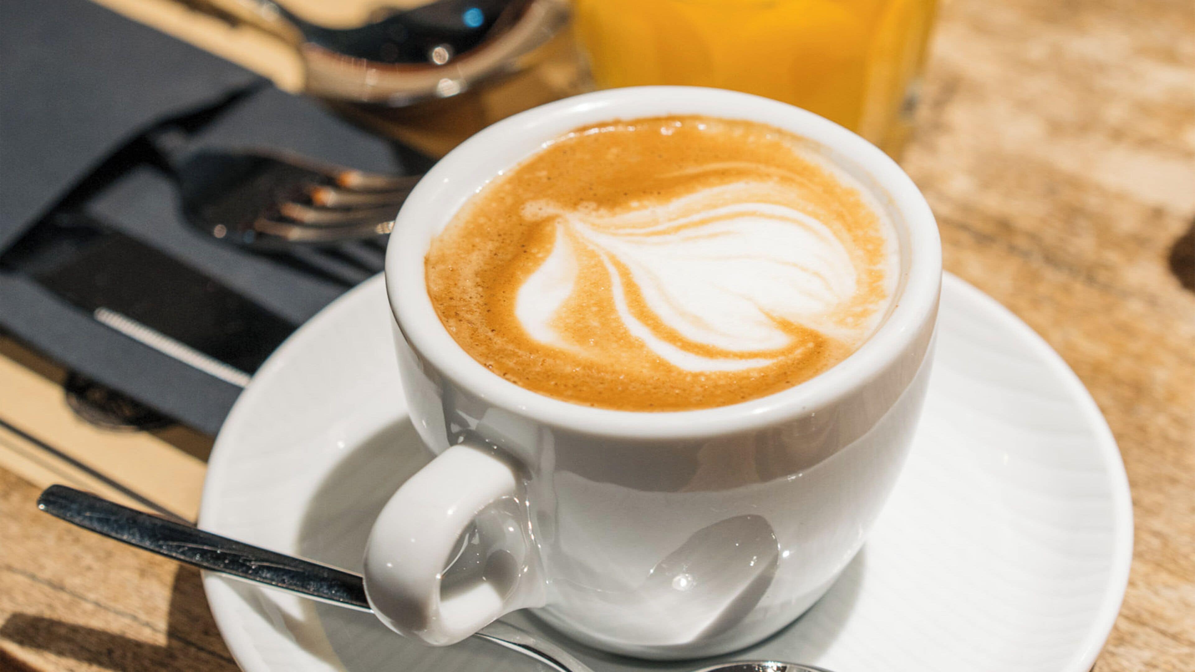 A cup of coffee with latte art on a saucer, a spoon, and a glass of orange juice on a table.