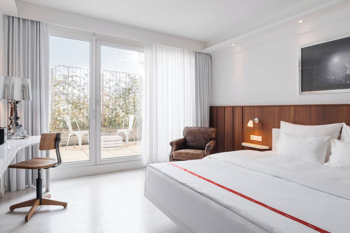 A hotel room with a bed, a desk with a chair, a small armchair, a nightstand with a lamp, and a large window with curtains.