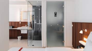 Una habitación de hotel moderna con cabina de ducha de vidrio, tocador de madera, cama y mesita de noche con lámpara.
