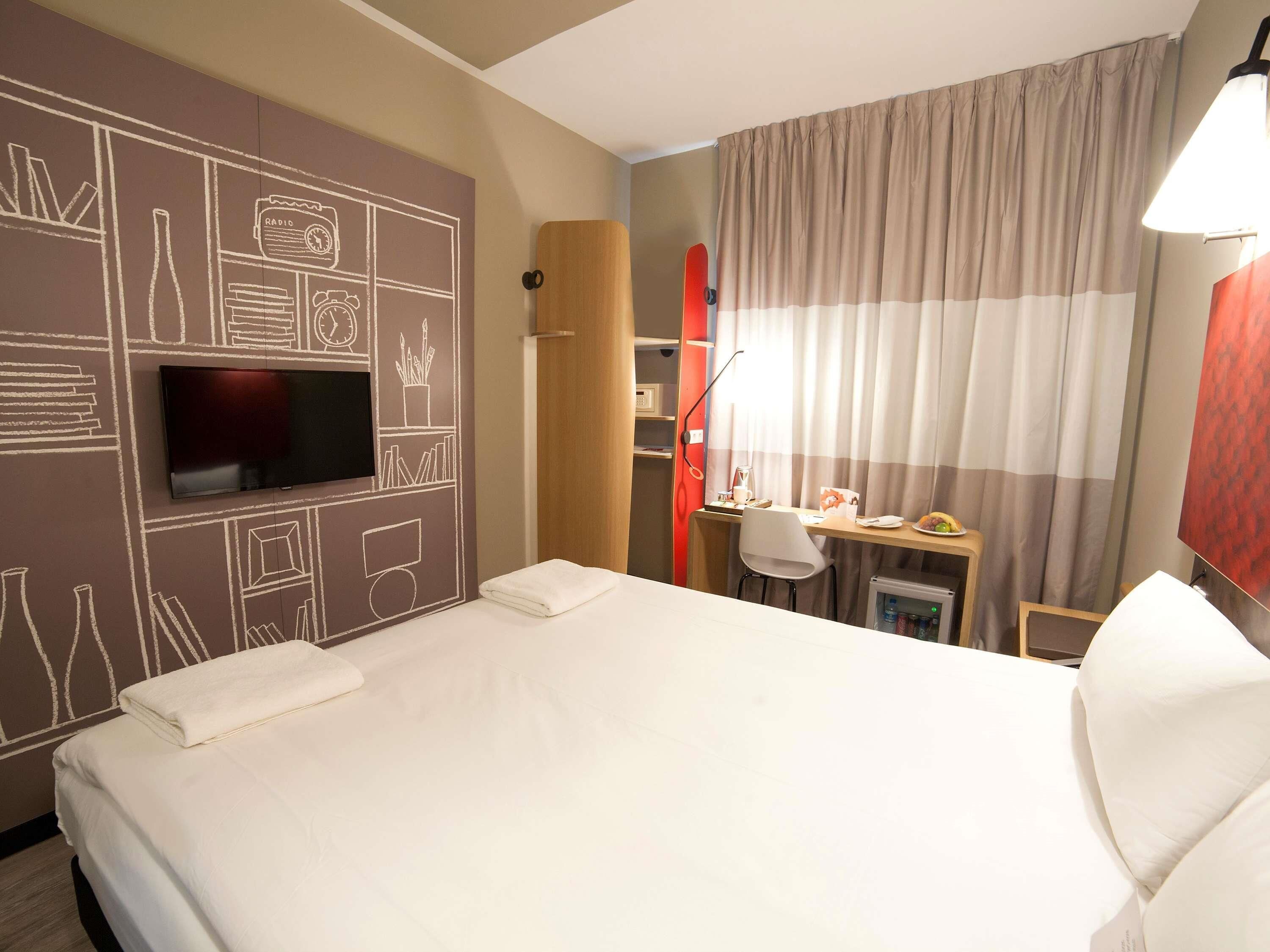 A hotel room with a bed, a TV, a desk with a chair, a small fridge, and curtains.