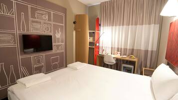 A hotel room with a bed, a TV, a desk with a chair, a small fridge, and curtains.