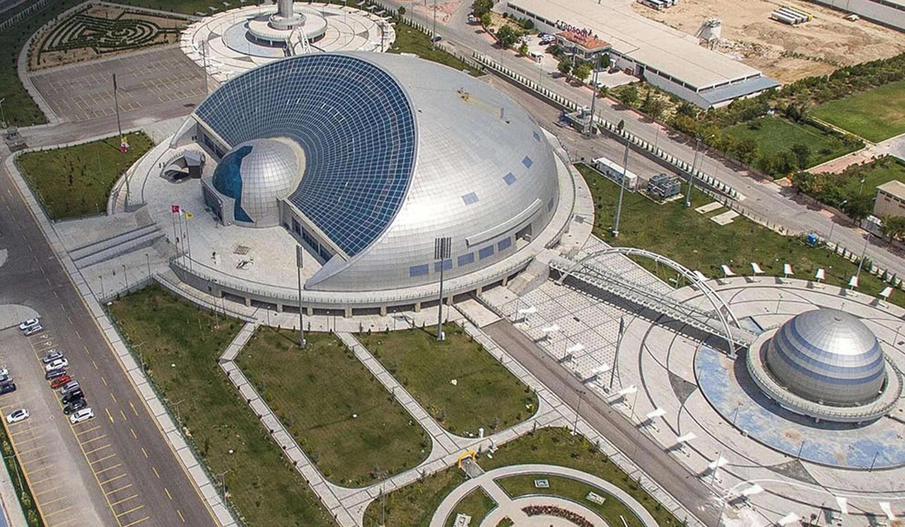 Un edificio grande y circular con una estructura de cúpula, rodeado de áreas ajardinadas y una carretera.