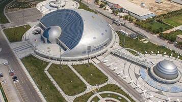 Un edificio grande y circular con una estructura de cúpula, rodeado de áreas ajardinadas y una carretera.