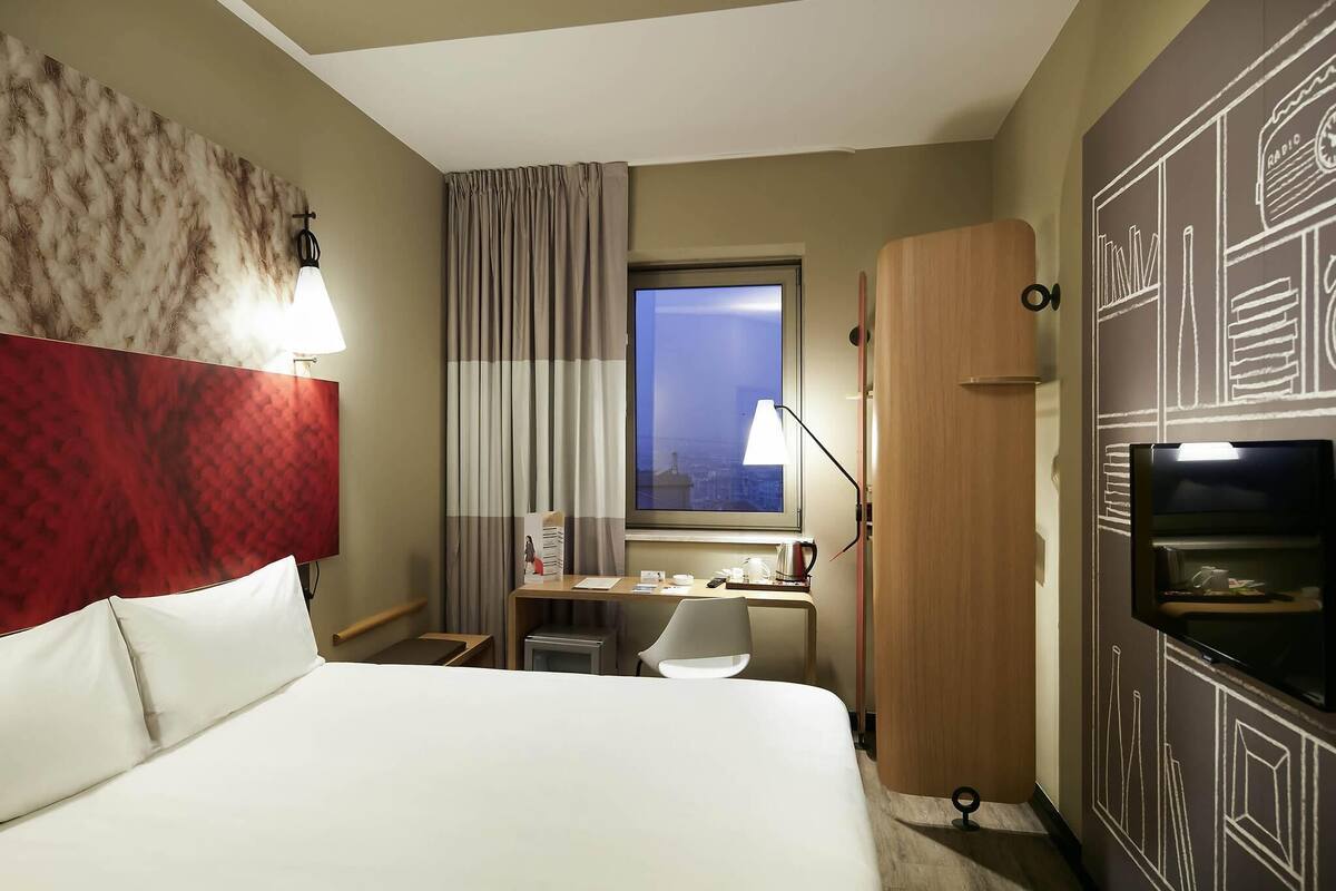 A hotel room with a bed, a desk, a chair, a window with curtains, a lamp, and a TV.