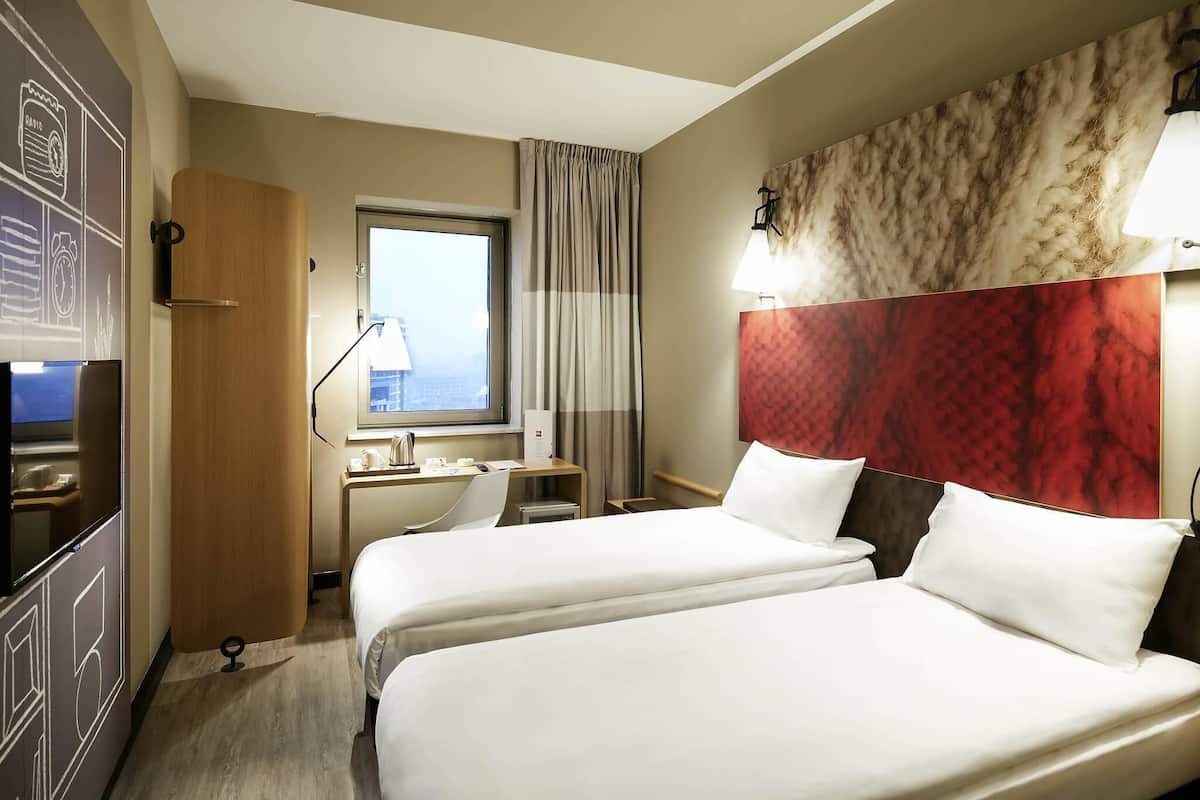 A hotel room with two beds, a desk, a chair, a window with curtains, and a wall with a textured design.
