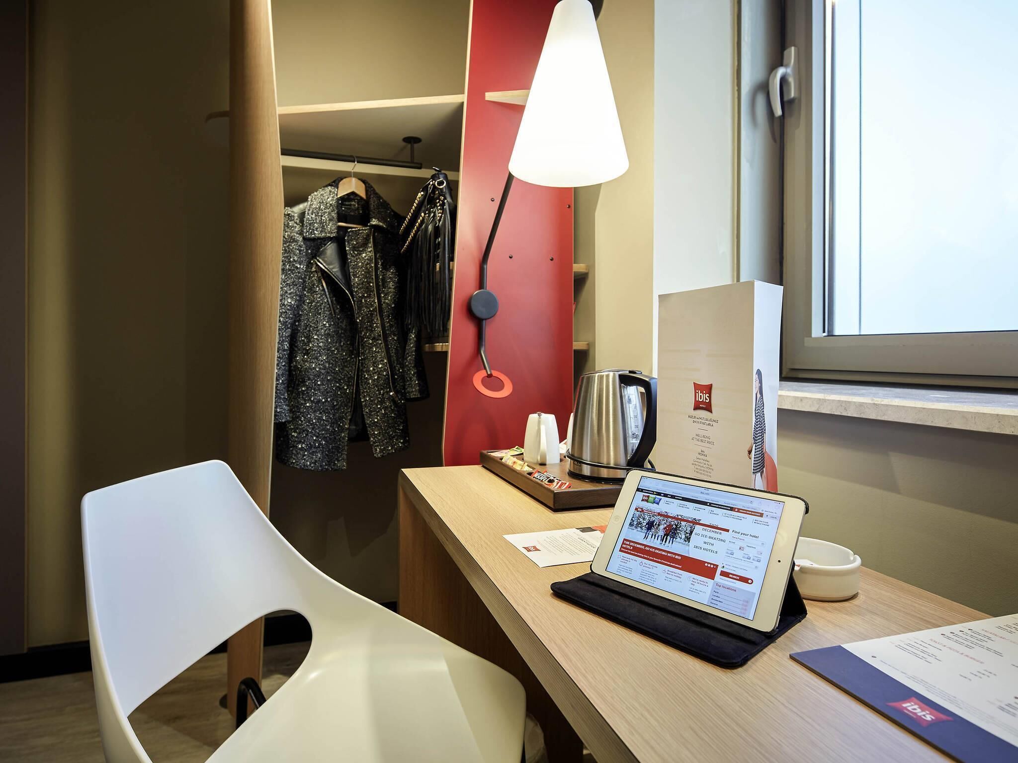 A hotel room with a desk, a laptop, a kettle, a coat rack with clothes, and a red wall.