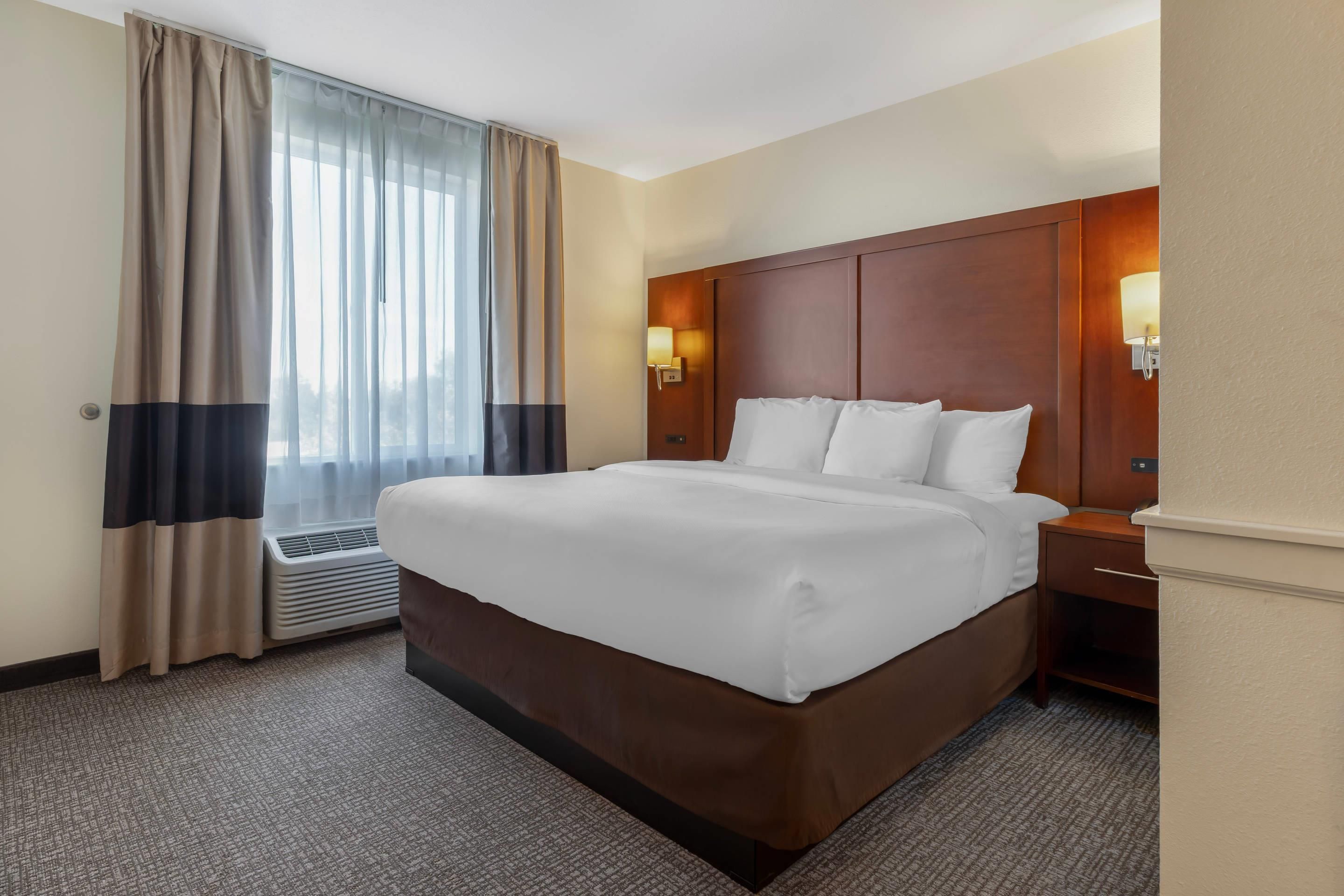 A hotel room with a large bed, a bedside table, a lamp, and a window with curtains.
