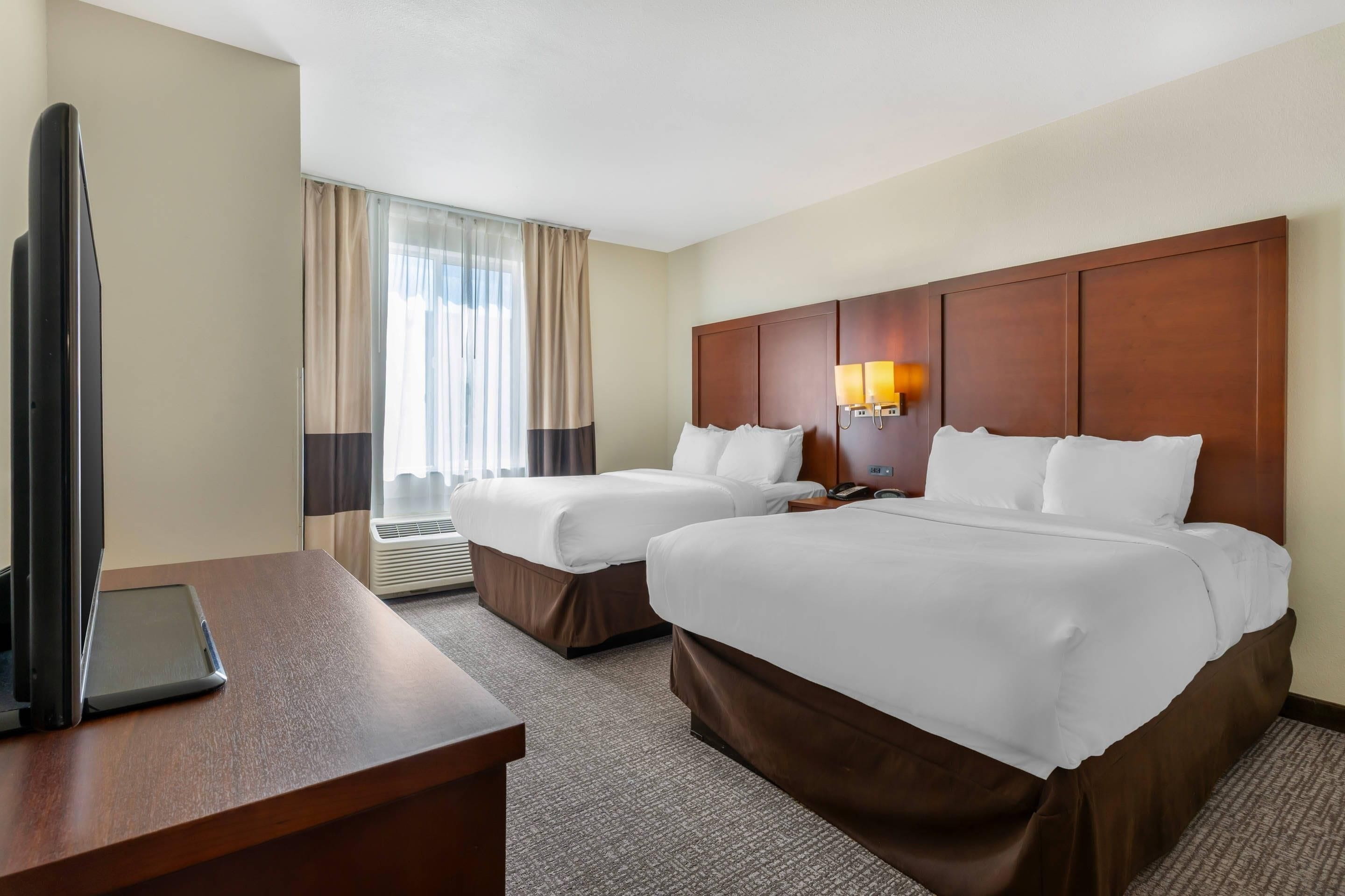 A hotel room with two beds, a desk, a TV, and an air conditioning unit.