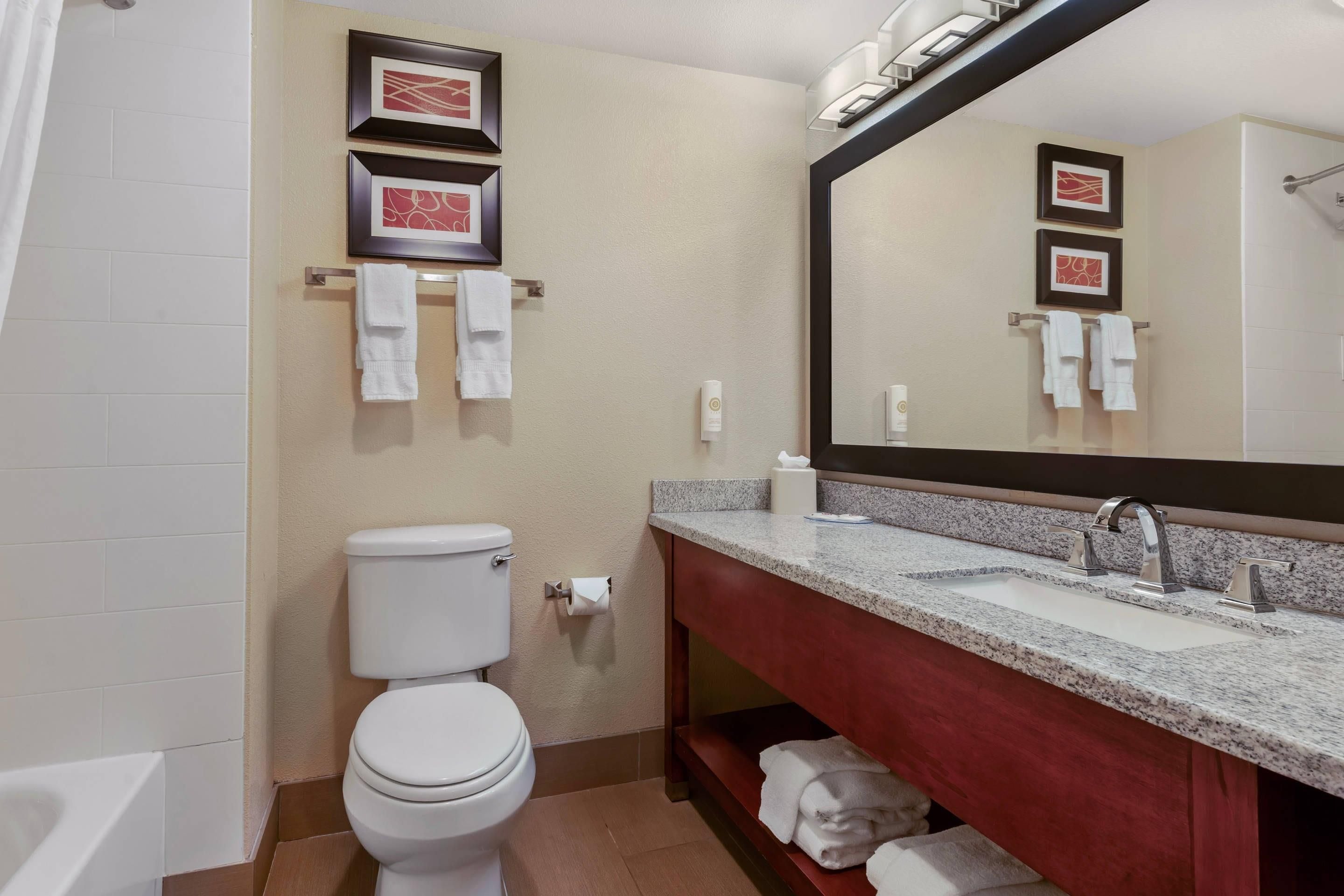 A bathroom with a toilet, sink, and mirror. There are towels hanging on a rack and a framed picture on the wall.