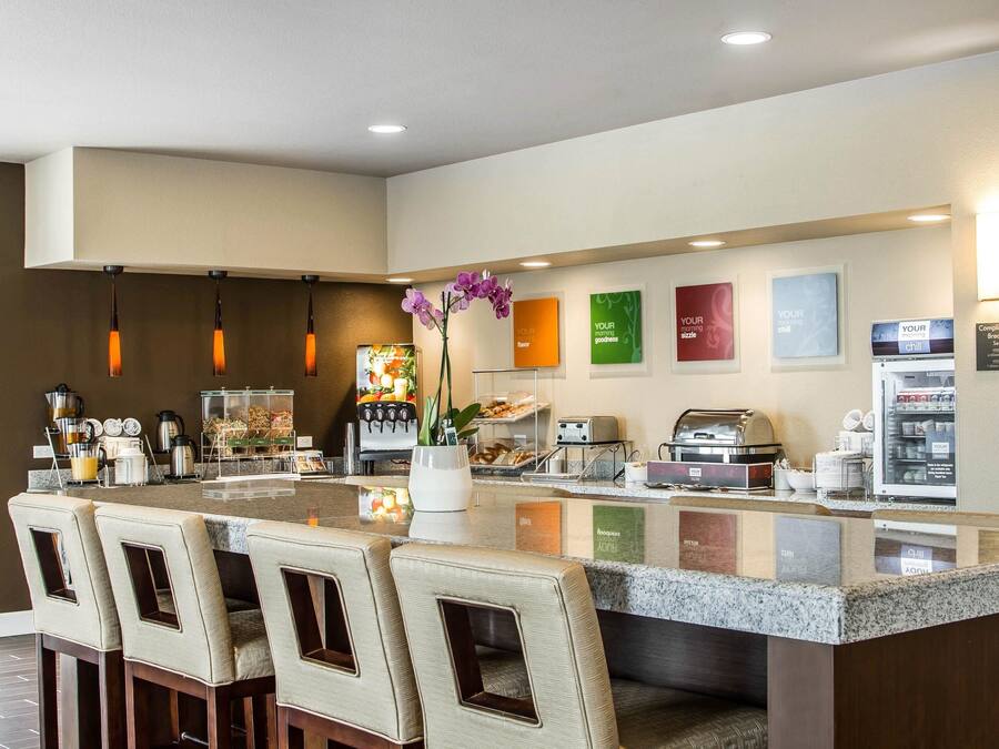 A hotel breakfast area with a central island, bar stools, a coffee machine, and a refrigerator.