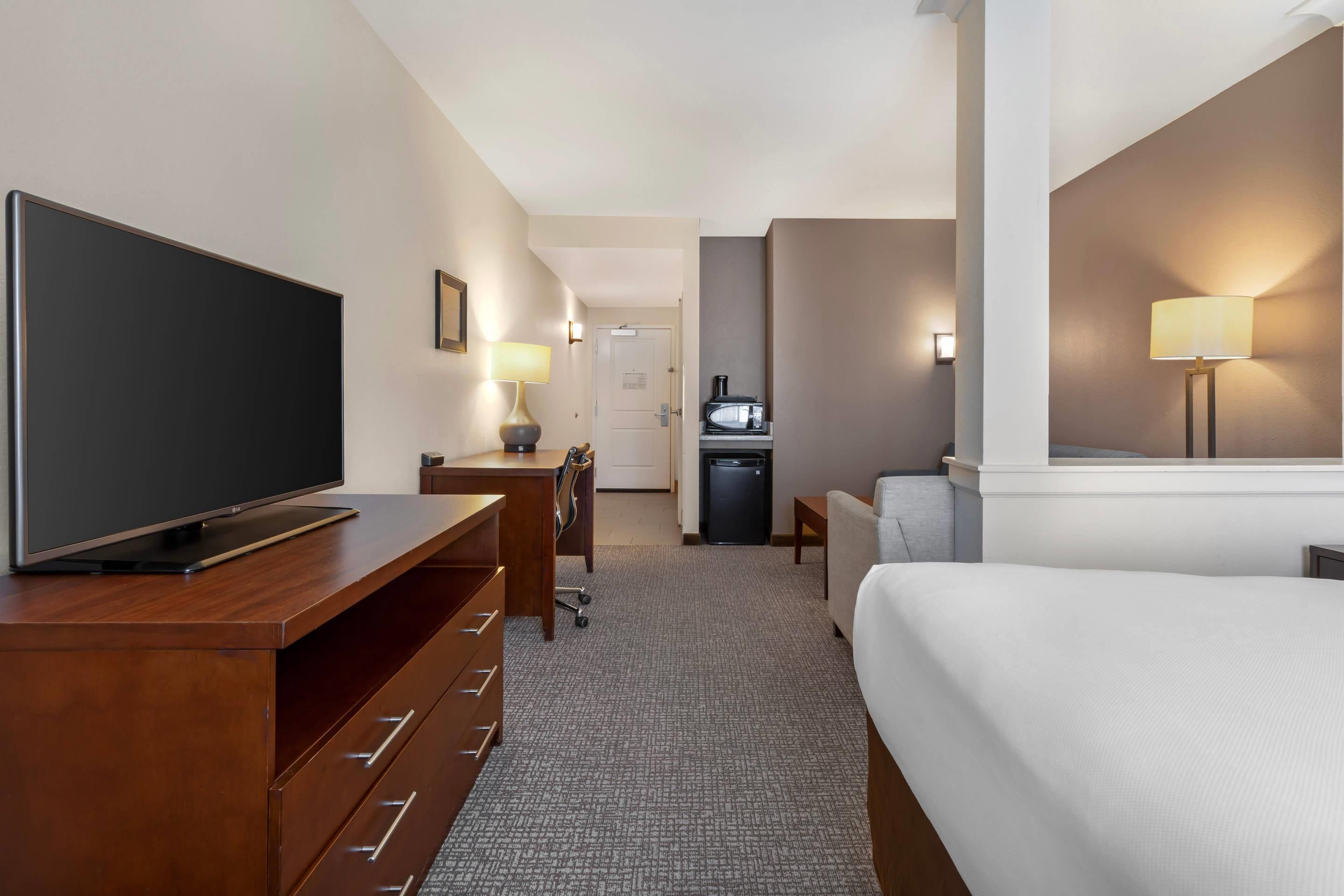 A hotel room with a flat-screen TV, a desk with a lamp, a bed, a small kitchenette, and a bathroom.