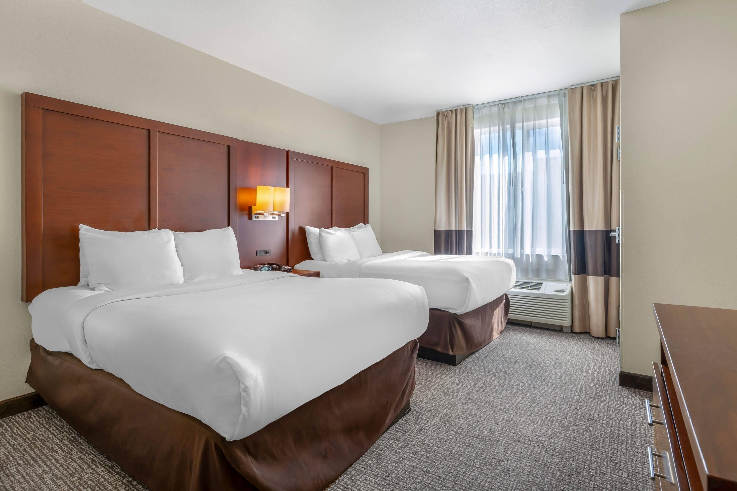 A hotel room with two beds, a nightstand, a lamp, and a window with curtains.