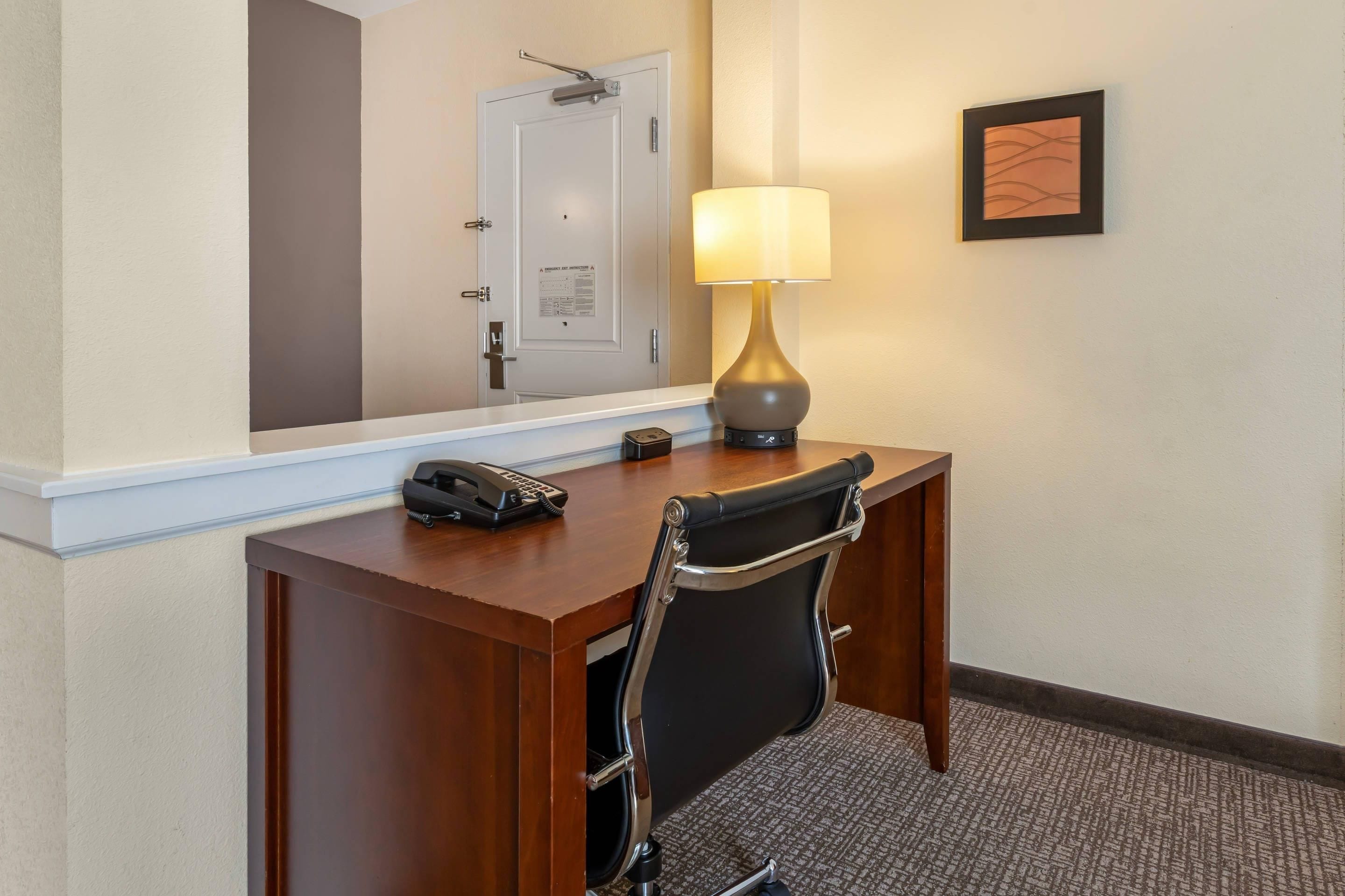Habitación de hotel con un escritorio de madera, un teléfono, una lámpara, una silla, un espejo y un cuadro en la pared.