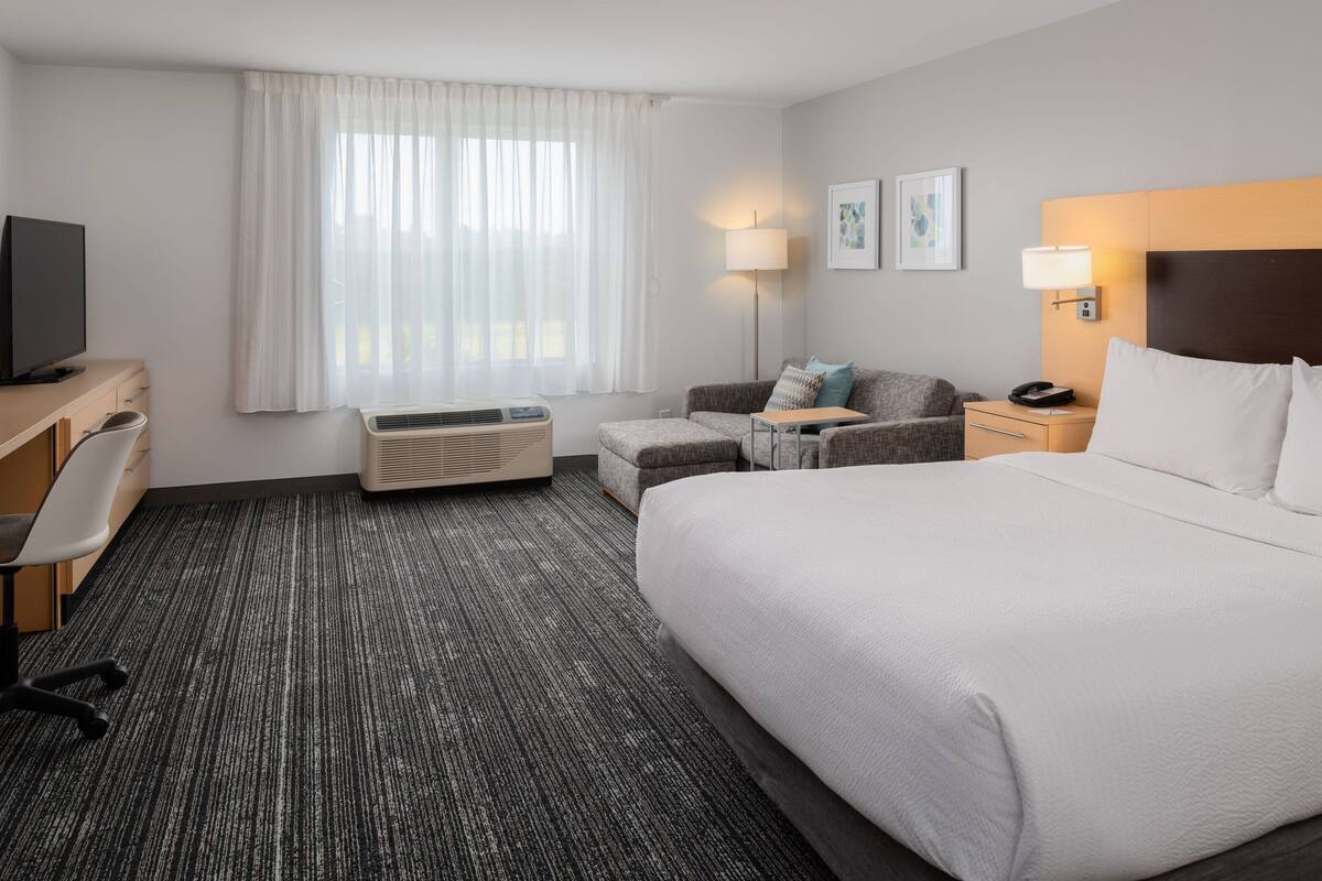 Une chambre d’hôtel avec un grand lit, un bureau avec un ordinateur, une chaise, un canapé, une télévision, une lampe et une fenêtre avec des rideaux.