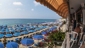 Vista sulla spiaggia con numerosi ombrelloni e lettini blu, un edificio in pietra con un balcone e una visuale libera sul mare.