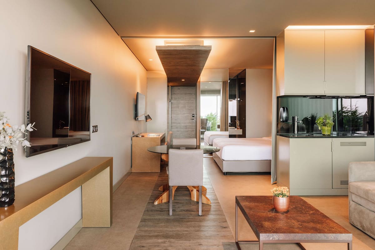 A modern hotel room with a sofa, a dining table, a kitchen area, and a large mirror.