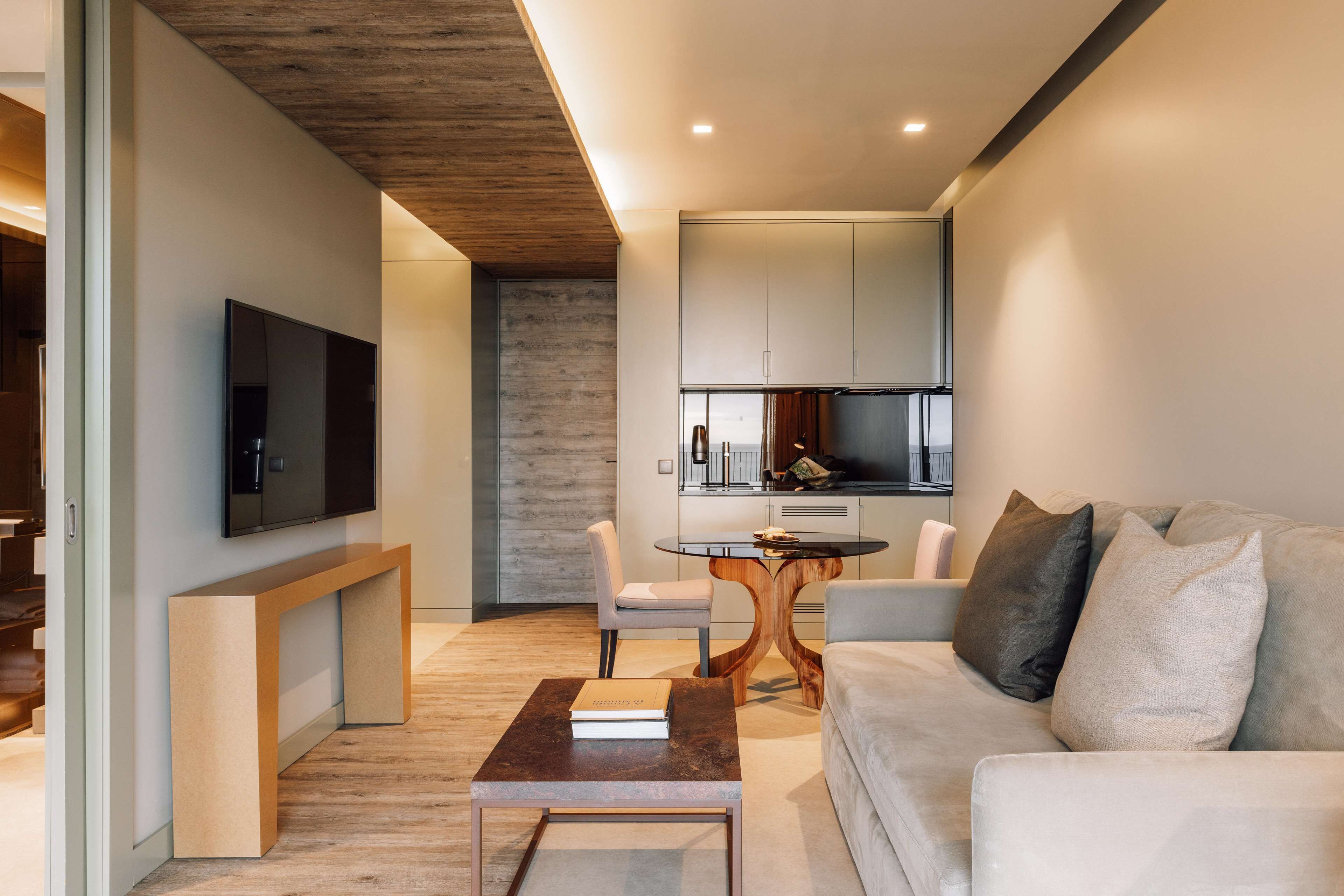 A modern living room with a sofa, coffee table, and TV. There is a dining area with a table and chairs. The kitchen is visible in the background.