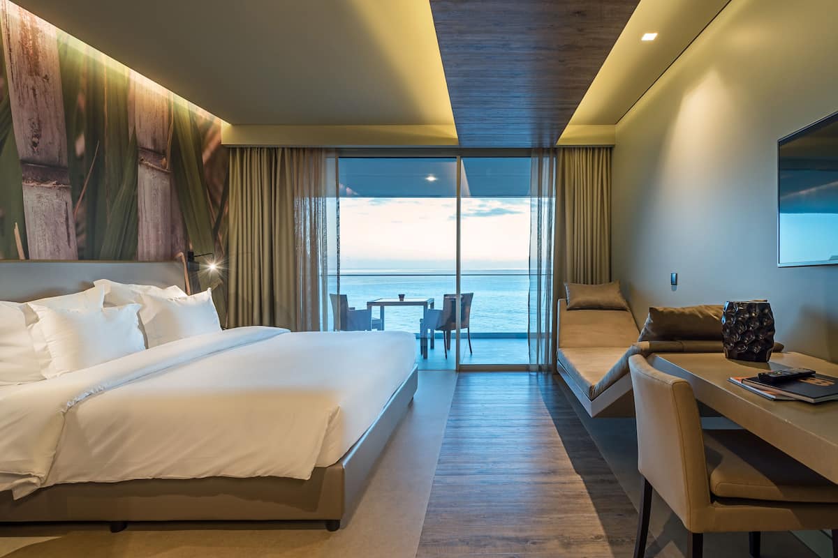 A hotel room with a large bed, a desk with a chair, and a view of the ocean through large windows.