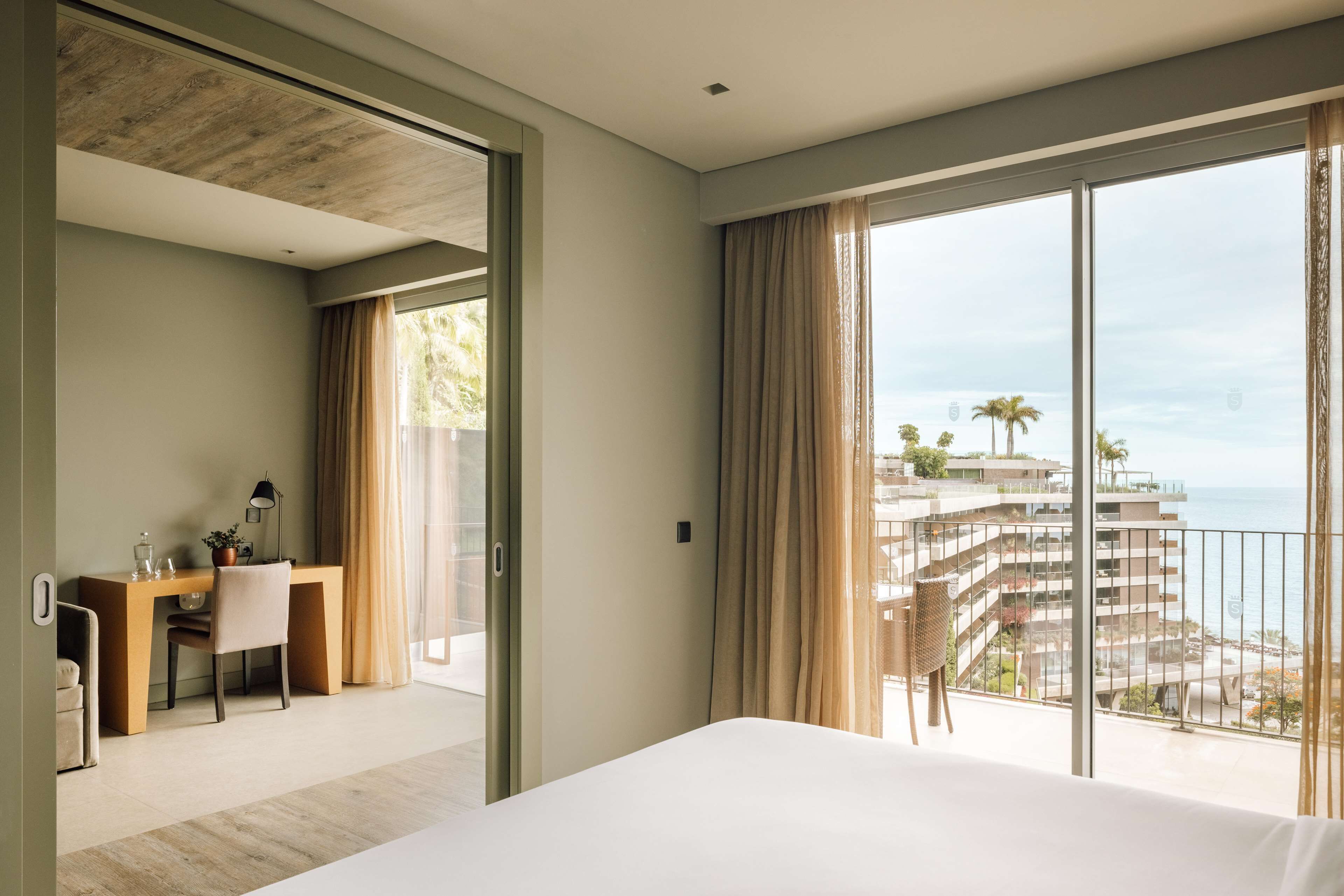A hotel room with a large bed, a desk with a chair, and a view of the ocean and buildings.